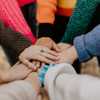 Eine Gruppe von Menschen steht im Kreis und legt ihre Hände übereinander in die Mitte. Sie tragen bunte Pullover in verschiedenen Farben wie Pink, Orange, Grün und Blau. 
