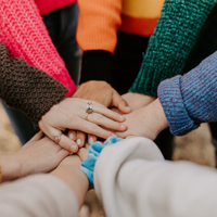 Eine Gruppe von Menschen steht im Kreis und legt ihre Hände übereinander in die Mitte. Sie tragen bunte Pullover in verschiedenen Farben wie Pink, Orange, Grün und Blau. 