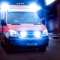 Ein Rettungswagen fährt mit eingeschaltetem Blaulicht und Sirene. Das Fahrzeug ist in den Farben Rot und Weiß gehalten und trägt die Aufschrift Rettungsdienst auf der Front. Die Scheinwerfer und das Blaulicht leuchten hell. 