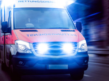 Ein Rettungswagen fährt mit eingeschaltetem Blaulicht und Sirene. Das Fahrzeug ist in den Farben Rot und Weiß gehalten und trägt die Aufschrift Rettungsdienst auf der Front. Die Scheinwerfer und das Blaulicht leuchten hell.