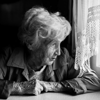 Eine ältere Frau mit welligem, grauem Haar sitzt an einem Tisch und schaut nachdenklich aus einem Fenster. Sie trägt eine Jacke und hat ihre Hände vor sich auf dem Tisch gefaltet. Das Licht, das durch das Fenster fällt, beleuchtet ihr Gesicht. Die Szene ist in Schwarz-Weiß gehalten.