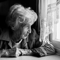 Eine ältere Frau mit welligem, grauem Haar sitzt an einem Tisch und schaut nachdenklich aus einem Fenster. Sie trägt eine Jacke und hat ihre Hände vor sich auf dem Tisch gefaltet. Das Licht, das durch das Fenster fällt, beleuchtet ihr Gesicht. Die Szene ist in Schwarz-Weiß gehalten.