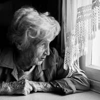 Eine ältere Frau mit welligem, grauem Haar sitzt an einem Tisch und schaut nachdenklich aus einem Fenster. Sie trägt eine Jacke und hat ihre Hände vor sich auf dem Tisch gefaltet. Das Licht, das durch das Fenster fällt, beleuchtet ihr Gesicht. Die Szene ist in Schwarz-Weiß gehalten.
