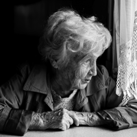 Eine ältere Frau mit welligem, grauem Haar sitzt an einem Tisch und schaut nachdenklich aus einem Fenster. Sie trägt eine Jacke und hat ihre Hände vor sich auf dem Tisch gefaltet. Das Licht, das durch das Fenster fällt, beleuchtet ihr Gesicht. Die Szene ist in Schwarz-Weiß gehalten.