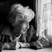 Eine ältere Frau mit welligem, grauem Haar sitzt an einem Tisch und schaut nachdenklich aus einem Fenster. Sie trägt eine Jacke und hat ihre Hände vor sich auf dem Tisch gefaltet. Das Licht, das durch das Fenster fällt, beleuchtet ihr Gesicht. Die Szene ist in Schwarz-Weiß gehalten.
