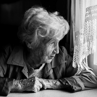 Eine ältere Frau mit welligem, grauem Haar sitzt an einem Tisch und schaut nachdenklich aus einem Fenster. Sie trägt eine Jacke und hat ihre Hände vor sich auf dem Tisch gefaltet. Das Licht, das durch das Fenster fällt, beleuchtet ihr Gesicht. Die Szene ist in Schwarz-Weiß gehalten.