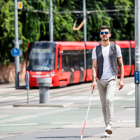 Ein junger blinder Mann läuft auf dem Gehweg. Er hat eine Sonnenbrille auf und einen Blindenstock in der Hand. Mit dem Blindenstock benutzt er die Leitstreifen auf dem Boden. Im Hintergrund fährt eine Straßenbahn entlang.
