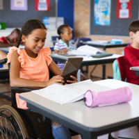 In einem Klassenzimmer sitzen mehrere Kinder an ihren Tischen und arbeiten konzentriert. Im Vordergrund sitzt ein Mädchen im Rollstuhl, das ein Tablet in den Händen hält und lächelt. Auf ihrem Tisch liegen Schulmaterialien wie Hefte und ein Federmäppchen. 