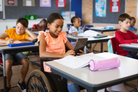 In einem Klassenzimmer sitzen mehrere Kinder an ihren Tischen und arbeiten konzentriert. Im Vordergrund sitzt ein Mädchen im Rollstuhl, das ein Tablet in den Händen hält und lächelt. Auf ihrem Tisch liegen Schulmaterialien wie Hefte und ein Federmäppchen. 