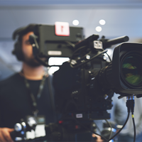 Ein Kameramann steht hinter einer professionellen Videokamera und richtet sie aus. Die Kamera ist groß und mit verschiedenen technischen Komponenten ausgestattet. Der Hintergrund ist unscharf, was darauf hindeutet, dass der Fokus auf der Kamera liegt. Der Kameramann trägt Kopfhörer und scheint sich auf die Aufnahme zu konzentrieren. Die Szene spielt sich in einem modernen, gut beleuchteten Innenraum ab.
