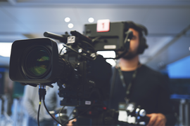 Ein Kameramann steht hinter einer professionellen Videokamera und richtet sie aus. Die Kamera ist groß und mit verschiedenen technischen Komponenten ausgestattet. Der Hintergrund ist unscharf, was darauf hindeutet, dass der Fokus auf der Kamera liegt. Der Kameramann trägt Kopfhörer und scheint sich auf die Aufnahme zu konzentrieren. Die Szene spielt sich in einem modernen, gut beleuchteten Innenraum ab.