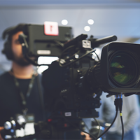 Ein Kameramann steht hinter einer professionellen Videokamera und richtet sie aus. Die Kamera ist groß und mit verschiedenen technischen Komponenten ausgestattet. Der Hintergrund ist unscharf, was darauf hindeutet, dass der Fokus auf der Kamera liegt. Der Kameramann trägt Kopfhörer und scheint sich auf die Aufnahme zu konzentrieren. Die Szene spielt sich in einem modernen, gut beleuchteten Innenraum ab.
