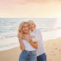 Das Bild zeigt zwei Frauen, die sich an einem Strand umarmen. Die jüngere Frau steht vorne und trägt ein weißes T-Shirt und eine blaue Jeans. Die ältere Frau steht hinter ihr und hat ihre Arme um die Schultern der jüngeren Frau gelegt. Beide lächeln glücklich in die Kamera. Im Hintergrund ist das Meer zu sehen.