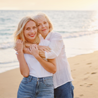 Das Bild zeigt zwei Frauen, die sich an einem Strand umarmen. Die jüngere Frau steht vorne und trägt ein weißes T-Shirt und eine blaue Jeans. Die ältere Frau steht hinter ihr und hat ihre Arme um die Schultern der jüngeren Frau gelegt. Beide lächeln glücklich in die Kamera. Im Hintergrund ist das Meer zu sehen.