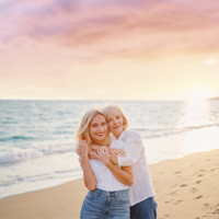 Das Bild zeigt zwei Frauen, die sich an einem Strand umarmen. Die jüngere Frau steht vorne und trägt ein weißes T-Shirt und eine blaue Jeans. Die ältere Frau steht hinter ihr und hat ihre Arme um die Schultern der jüngeren Frau gelegt. Beide lächeln glücklich in die Kamera. Im Hintergrund ist das Meer zu sehen.