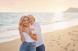 Das Bild zeigt zwei Frauen, die sich an einem Strand umarmen. Die jüngere Frau steht vorne und trägt ein weißes T-Shirt und eine blaue Jeans. Die ältere Frau steht hinter ihr und hat ihre Arme um die Schultern der jüngeren Frau gelegt. Beide lächeln glücklich in die Kamera. Im Hintergrund ist das Meer zu sehen.