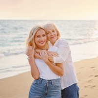 Das Bild zeigt zwei Frauen, die sich an einem Strand umarmen. Die jüngere Frau steht vorne und trägt ein weißes T-Shirt und eine blaue Jeans. Die ältere Frau steht hinter ihr und hat ihre Arme um die Schultern der jüngeren Frau gelegt. Beide lächeln glücklich in die Kamera. Im Hintergrund ist das Meer zu sehen.