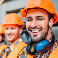 Das Bild zeigt drei Bauarbeiter, die Schutzhelme und reflektierende Westen tragen. Der Arbeiter im Vordergrund lächelt in die Kamera und trägt zusätzlich Gehörschutz um den Hals. Die beiden anderen Arbeiter im Hintergrund sind ebenfalls in Arbeitskleidung. Im Hintergrund ist eine Baustelle zu erkennen.