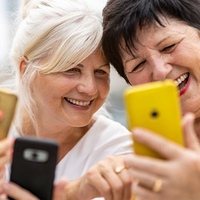 Drei ältere Frauen sitzen zusammen und lachen, während sie auf ihre Smartphones schauen. Alle drei halten ihre Telefone in den Händen und wirken entspannt und glücklich. Die Frau links trägt eine Brille und ein hellblaues Oberteil, die Frau in der Mitte hat weißes Haar und trägt ein weißes Oberteil, und die Frau rechts hat dunkles Haar und trägt ein weißes Oberteil.