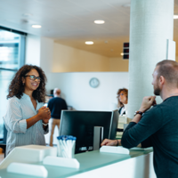 In einem modernen, hellen Bürogebäude stehen drei Personen an einem Empfangstresen. Eine Frau mit lockigem Haar und Brille steht hinter dem Tresen und lächelt freundlich. Vor dem Tresen stehen ein Mann und eine Frau, die beide ebenfalls lächeln. Im Hintergrund sind weitere Personen zu sehen, die sich in der offenen und gut beleuchteten Umgebung bewegen. Pflanzen und moderne Möbel tragen zur angenehmen Atmosphäre bei.