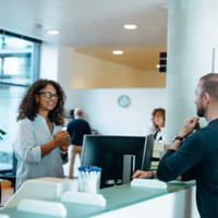 In einem modernen, hellen Bürogebäude stehen drei Personen an einem Empfangstresen. Eine Frau mit lockigem Haar und Brille steht hinter dem Tresen und lächelt freundlich. Vor dem Tresen stehen ein Mann und eine Frau, die beide ebenfalls lächeln. Im Hintergrund sind weitere Personen zu sehen, die sich in der offenen und gut beleuchteten Umgebung bewegen. Pflanzen und moderne Möbel tragen zur angenehmen Atmosphäre bei.