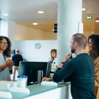 In einem modernen, hellen Bürogebäude stehen drei Personen an einem Empfangstresen. Eine Frau mit lockigem Haar und Brille steht hinter dem Tresen und lächelt freundlich. Vor dem Tresen stehen ein Mann und eine Frau, die beide ebenfalls lächeln. Im Hintergrund sind weitere Personen zu sehen, die sich in der offenen und gut beleuchteten Umgebung bewegen. Pflanzen und moderne Möbel tragen zur angenehmen Atmosphäre bei.