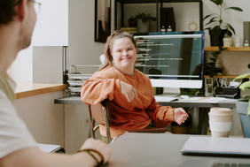 Eine junge Frau sitzt in einem Büro vor einem Computerbildschirm und spricht mit einer anderen Person im Vordergrund. Sie trägt einen orangefarbenen Pullover, lächelt und gestikuliert mit der Hand. Auf dem Schreibtisch stehen unter anderem ein Kaffeebecher und technische Geräte.