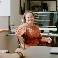 Eine junge Frau sitzt in einem Büro vor einem Computerbildschirm und spricht mit einer anderen Person im Vordergrund. Sie trägt einen orangefarbenen Pullover, lächelt und gestikuliert mit der Hand. Auf dem Schreibtisch stehen unter anderem ein Kaffeebecher und technische Geräte.