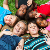 Auf dem Bild liegen sechs Kinder auf dem Rasen in einem Kreis und lächeln in die Kamera. Sie haben ihre Köpfe nah beieinander, sodass sie einen geschlossenen Kreis bilden. Die Kinder tragen bunte Sommerkleidung und scheinen fröhlich und entspannt zu sein. Einige von ihnen winken der Kamera zu. Der Hintergrund besteht aus grünem Gras.
