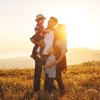 Eine Familie steht auf einer Wiese bei Sonnenuntergang. Ein Mann hält ein kleines Kind im Arm, während eine Frau und ein weiteres Kind neben ihnen stehen. Alle lächeln. Im Hintergrund erstreckt sich eine Landschaft mit Hügeln und einem Gewässer.