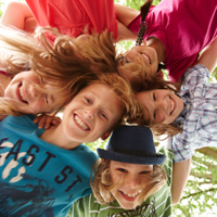 Auf dem Bild sind sechs fröhliche Kinder zu sehen, die in einem Kreis stehen und ihre Köpfe zusammenstecken. Sie lachen und schauen nach unten in die Kamera. Die Kinder tragen bunte Kleidung. Im Hintergrund sind grüne Blätter von Bäumen zu erkennen. 