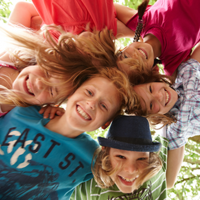 Auf dem Bild sind sechs fröhliche Kinder zu sehen, die in einem Kreis stehen und ihre Köpfe zusammenstecken. Sie lachen und schauen nach unten in die Kamera. Die Kinder tragen bunte Kleidung. Im Hintergrund sind grüne Blätter von Bäumen zu erkennen. 