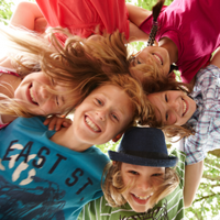 Auf dem Bild sind sechs fröhliche Kinder zu sehen, die in einem Kreis stehen und ihre Köpfe zusammenstecken. Sie lachen und schauen nach unten in die Kamera. Die Kinder tragen bunte Kleidung. Im Hintergrund sind grüne Blätter von Bäumen zu erkennen. 