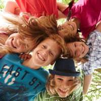 Auf dem Bild sind sechs fröhliche Kinder zu sehen, die in einem Kreis stehen und ihre Köpfe zusammenstecken. Sie lachen und schauen nach unten in die Kamera. Die Kinder tragen bunte Kleidung. Im Hintergrund sind grüne Blätter von Bäumen zu erkennen. 